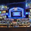 The image shows an audience seated in a theater with a stage set for a performance, viewed from a balcony level with refreshments in the foreground.