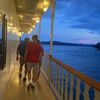 Two people are walking on the deck of a ship at dusk, with a view of the water and a colorful sky in the background.