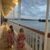 Two children in matching floral dresses are walking along a covered waterside gallery with ornate railings, as evening light reflects off the wet ground and a tranquil body of water sits in the background beneath a cloudy sky.