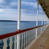 The image shows the perspective view of an outdoor corridor on a boat with balconies on the right-hand side, overlooking a calm body of water with a distant boat and treelined coast under an overcast sky.