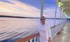 A person is standing on the deck of a ship, smiling for the camera, with a serene sunset over calm waters in the background.