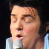 A performer dressed as Elvis Presley is singing on stage with band members and backup singers in the background.