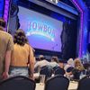 Audience members are seated at tables facing an ornately framed stage with the words 