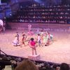 A group of performers dressed in traditional Western attire are engaging in a theatrical scene in a sand-covered arena, while a large audience watches from surrounding tiered seating.