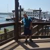 A person is posing with a smile on a wooden deck with a large multi-deck boat docked in the background.