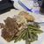 The image shows a plate of food consisting of roast beef, mashed potatoes with gravy, and green beans, with a beverage and an empty plate in the background on a table with a blue napkin.