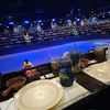 The image shows a dining area set with plates and drinks, overlooking an indoor arena with a large audience seated in preparation for an event or performance.