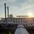 The image shows a moored paddle-wheel steamboat at sunset with the sun low in the sky behind it, casting a warm glow over the scene.