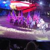 Riders on horseback carry striped flags in a patriotic performance, illuminated under theatrical lighting with an audience in the foreground.