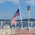 An American flag flutters in the breeze on a ferry deck with white chairs and a lamp post, against a backdrop of a calm lake and hills under a blue sky.