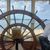 A person is at the helm of a boat, steering with a large wooden ship's wheel with a scenic view of the water and landscape ahead.