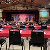 The image shows a dinner theater setup with numbered tables, black chairs, red tablecloths, and a stage with a rustic set design and lighting equipment overhead.