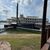 The image presents a large, multi-deck riverboat with iconic paddlewheel elements, docked beside a grassy waterfront under a partly cloudy sky.