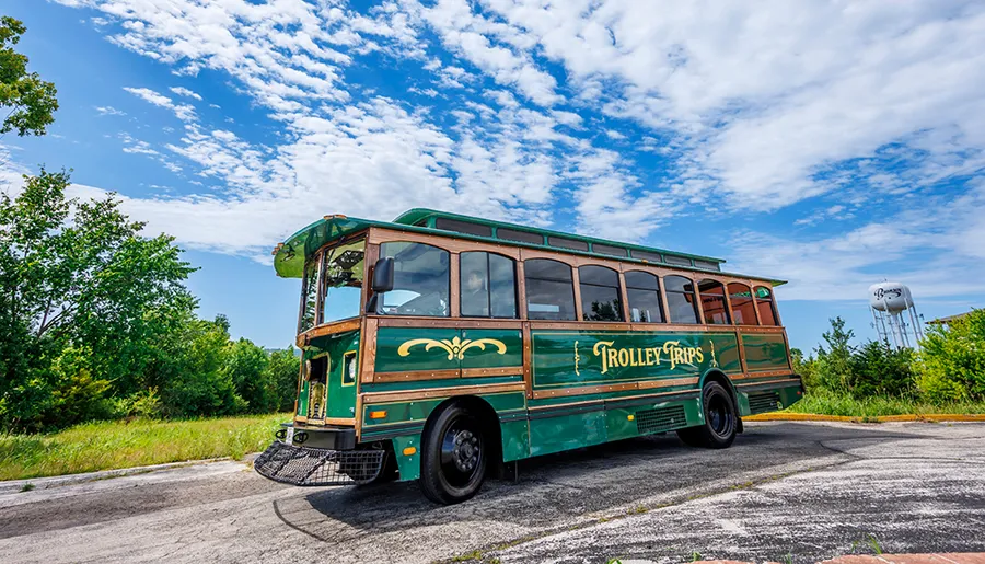 A green trolley-style bus with Trolley Trips written on its side is parked under a blue sky dotted with clouds, beside a roadside with surrounding vegetation and a water tower in the background.