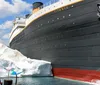 This image features a group of people looking at a large stationary replica of the Titanic ship next to an artificial iceberg likely part of a themed attraction or exhibit