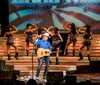 A performer in a cowboy hat is exuberantly singing on stage with musicians and dancers in the background all set against an oversized clock backdrop