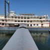 A large, multi-deck paddle steamer with a red paddle wheel is docked at the end of a wooden pier on a calm body of water under a clear sky.