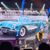 A presenter is speaking on stage with a backdrop featuring a 1959 Corvette and text about the launch of the Barbie doll, illuminated by stage lights and accompanied by musicians.