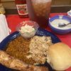 A hearty meal consisting of a grilled pork rib, shredded meat, baked beans, coleslaw, a bread roll, alongside a bowl of cream soup and a glass of iced tea, with condiments like ketchup and mustard present on the table.