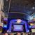 The image depicts the interior of a lavish theater with an audience waiting for a performance beneath a sign reading 