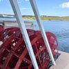 The image shows the side view of a red paddle wheel of a boat, with a backdrop of a blue sky and a distant shoreline.