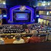 The image shows an audience seated at tables inside a venue, looking towards a stage with a decorative arch that reads 