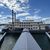 The image displays a large, multi-deck riverboat with a traditional paddlewheel, docked at a pier with a walkway leading up to it under a partly cloudy sky.