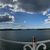 The image captures a panoramic view of a serene lake with a glittering sun above, clouds scattered across the sky, and a partial view of a person beside a white fence.