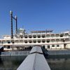 The image shows a large, multi-decked riverboat with traditional paddle-wheel design, moored at a dock with a long gangway on a clear day.