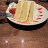 A slice of layered vanilla cake with white frosting, garnished with a strawberry and decorative sauce, is served on a white plate with a fork on the side, accompanied by a cup and glass on a wooden table.