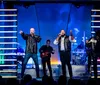 Four singers are energetically performing on stage with a band in the background illuminated by colorful stage lighting