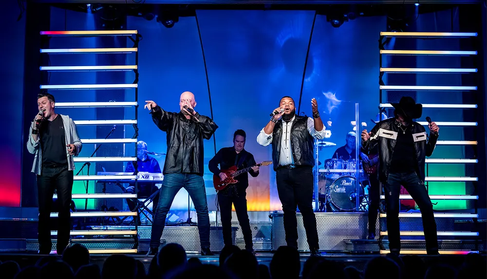 Four singers are energetically performing on stage with a band in the background illuminated by colorful stage lighting