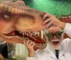 A person in a lab coat and goggles is playfully posing with a large model of a dinosaurs head