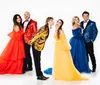 Five people wearing colorful formal attire pose in front of a white background with two couples on the sides and a man in the center playfully pouting towards a woman in yellow