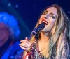 A woman is passionately singing into a microphone on stage with blurred stage lights in the background and a musician partially visible behind her