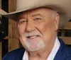 A man with a white mustache and beard is wearing a large white cowboy hat and a blue jacket posing in front of a wooden background