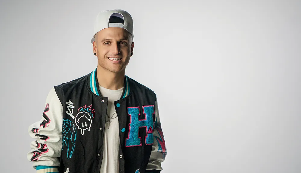 A smiling man wearing a trendy varsity jacket and a baseball cap poses against a light gray background