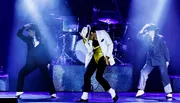 Three performers onstage exhibit a dynamic dance pose reminiscent of Michael Jackson's iconic style, with the central figure donning a white jacket and gold belt.