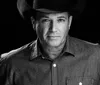 The image depicts a black and white portrait of a man wearing a cowboy hat and a patterned shirt gazing directly at the camera against a black background