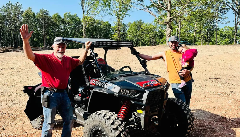 Two smiling men and a child standing next to an off-road vehicle in a sunny outdoor setting with one man waving at the camera