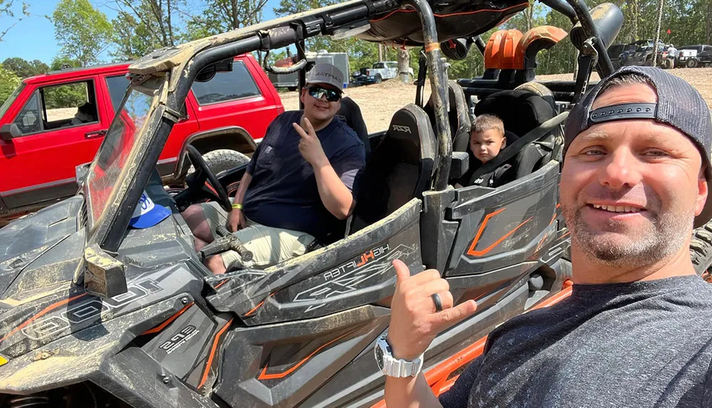 Three people are posing for a photo with a man in the foreground showing a thumbs-up and the others sitting in a dirt-covered off-road vehicle with a red SUV in the background