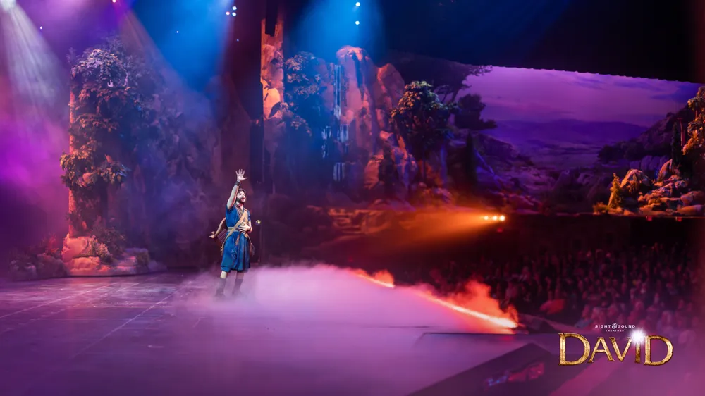 A performer in historical costume stands on a foggy stage with dramatic lighting as part of a theatrical production set in a natural landscape while the audience watches