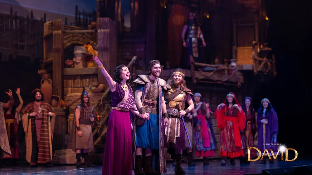 The image depicts a lively scene from a stage production with performers in vibrant period costumes appearing to be in mid-song or dialogue set against an elaborate set that suggests a historical or fantasy context