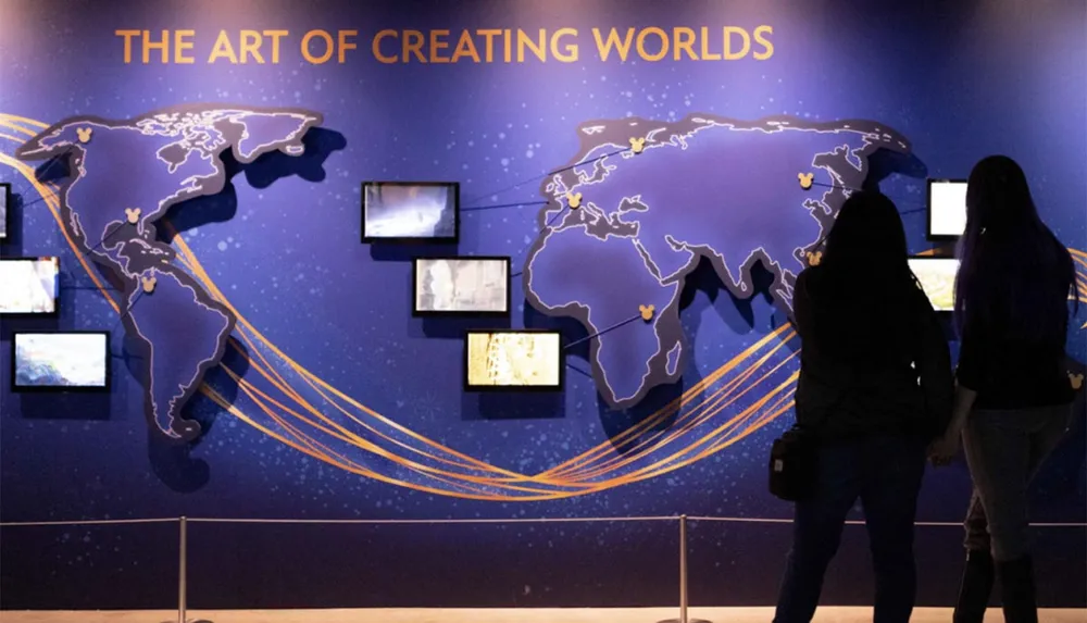 Two people are observing an exhibit titled THE ART OF CREATING WORLDS with world map graphics and multiple screens displaying content on a gallery wall