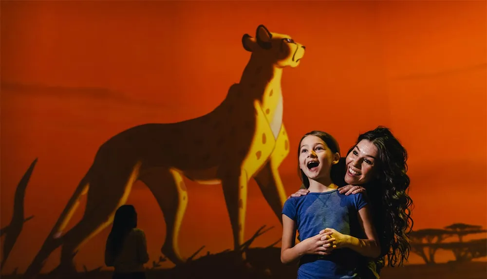 A woman and a young girl pose with bright smiles in front of a large projected animation of a cheetah against an orange backdrop