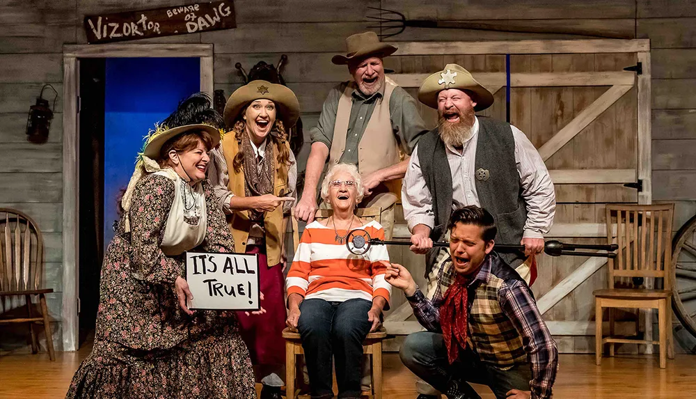 A group of costumed actors is posing enthusiastically on a theater set that evokes a wild west theme with one actor holding a sign that reads ITS ALL TRUE