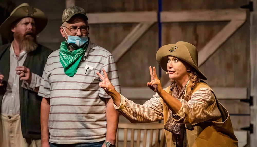 Three individuals in what appears to be a theatrical setting engage in lively interaction with the foremost person dressed in old-western attire gesturing animatedly