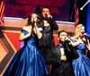 A group of performers in elegant blue gowns and sparkly suits sing into microphones on a brightly lit stage