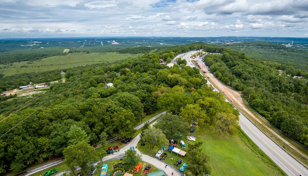 This image captures an aerial view of a curved highway slicing through a lush green landscape with clusters of vehicles and infrastructure dotted alongside