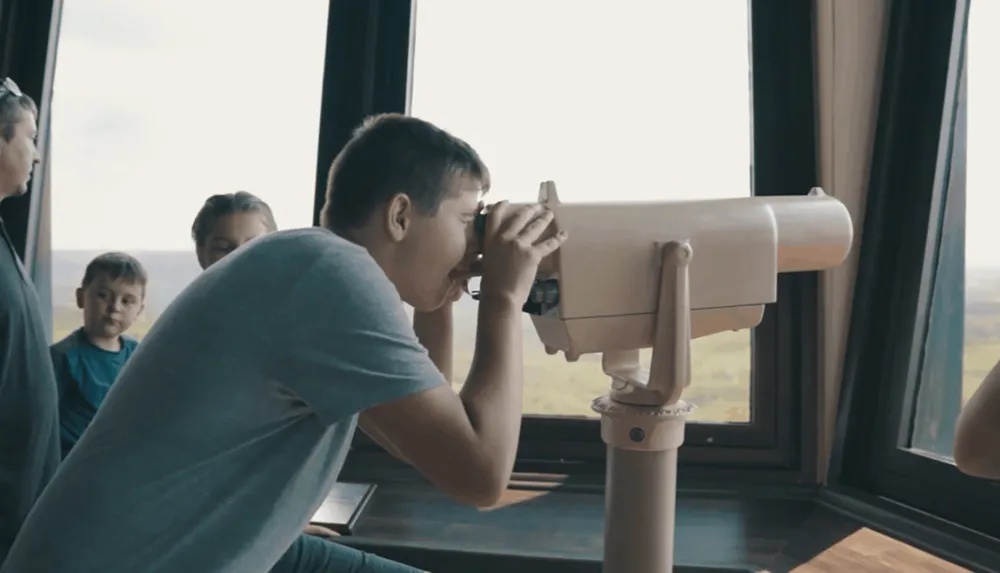 A person is looking through a stationary telescope with others waiting around possibly at a scenic viewpoint or observation deck